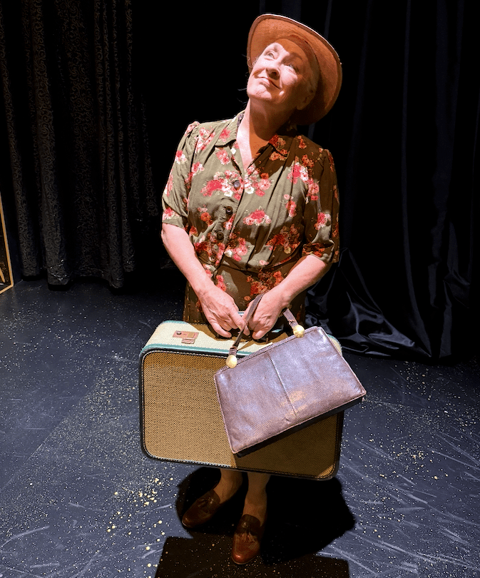 Woman in hat and dress holding purse and suitcase under a spotlight on a stage.
