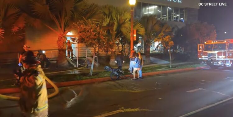 Fire trucks and firefighters work at night with hoses running into a door. A couple people are standing around a couple bikes lying on their sides on the curb. OC Street Live watermark.