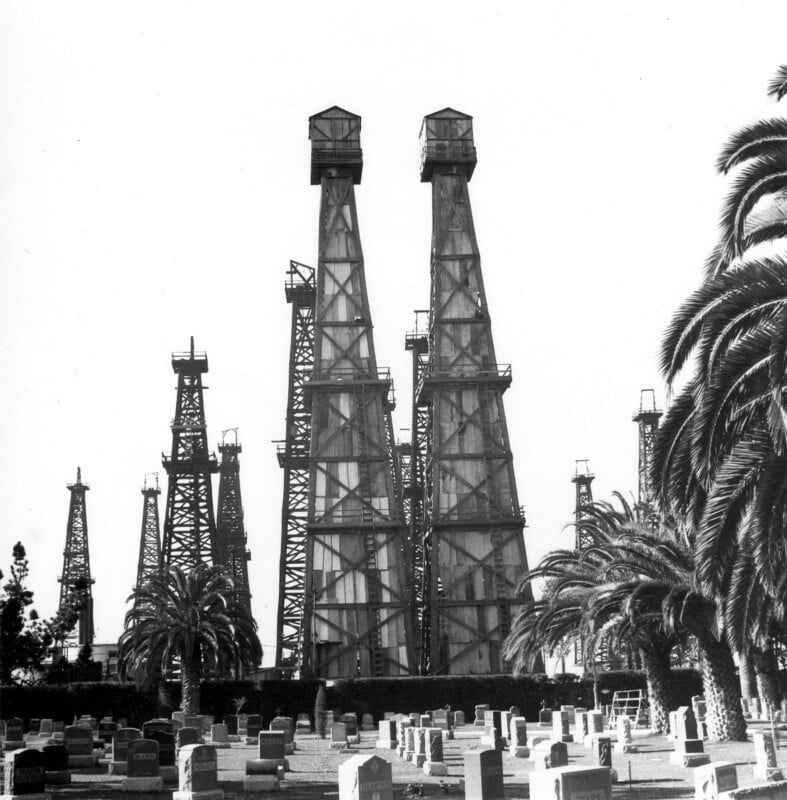 Black and white photo of several tall oil derricks behind a cemetery with rows of headstones and palm trees in the foreground.