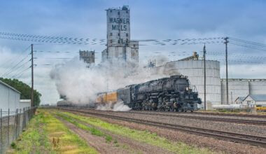 Big Boy steam locomotive and train passing a grain elevator in a rural town.