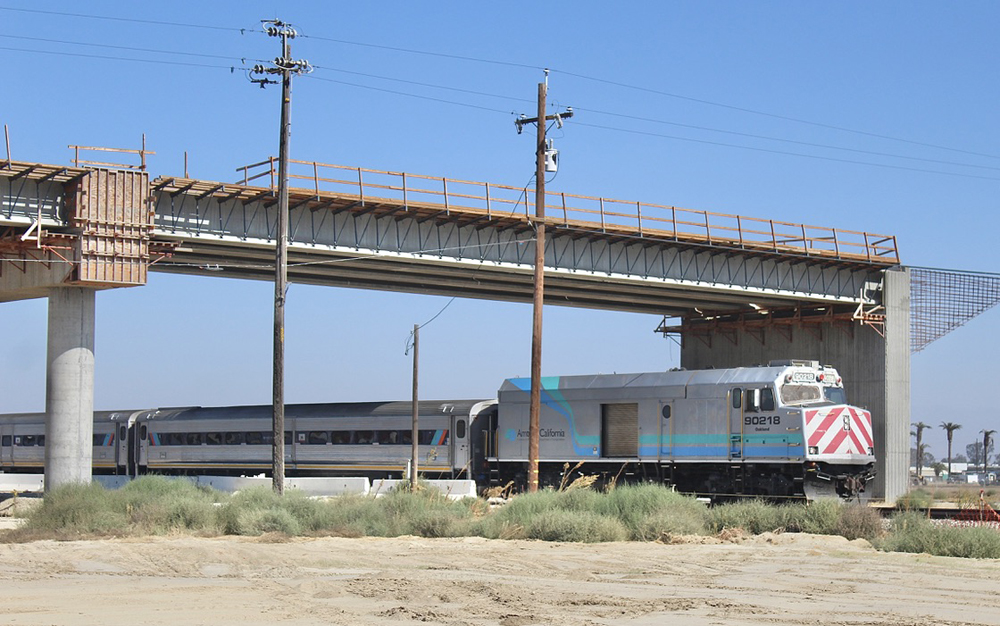 Passenger train travels beneath bridge under construction.