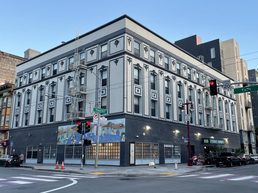 A corner building with geometric facade designs stands at an intersection. Traffic lights are visible, and there's a mural on the ground floor. Cones and street signs are present.