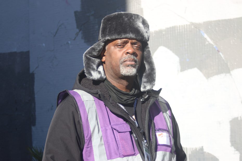 A man wearing a black winter hat and a purple safety vest stands outdoors in front of a gray and white painted wall.