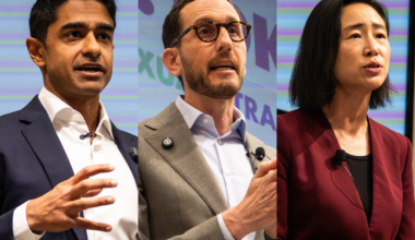 Three people stand side by side, each speaking or gesturing during a public event or debate, dressed in business attire against a mixed color background.