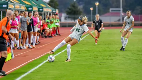 Julia Lucero A woman with blonde hair in a white and dark green football kit. She is on a football field and is in motion kicking the ball. One of her arms is in a white cast