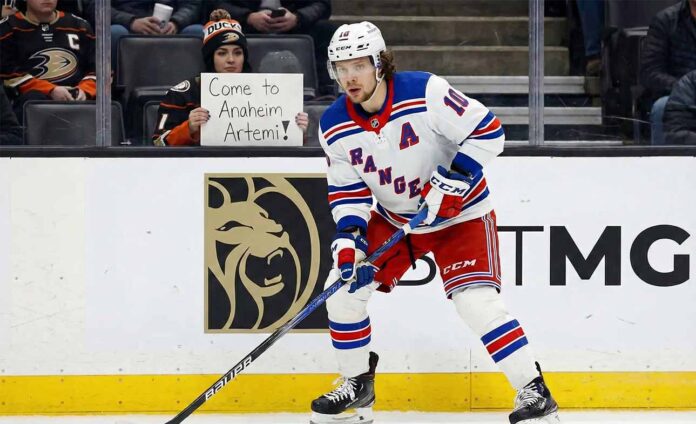 artemi-panarin-rangers-trade-rumors-ducks Artemi Panarin, wearing a white New York Rangers away jersey with a blue 'A', is on the ice with his stick during a game. In the background, a fan holds a sign that reads, "Come to Anaheim Artemi!".