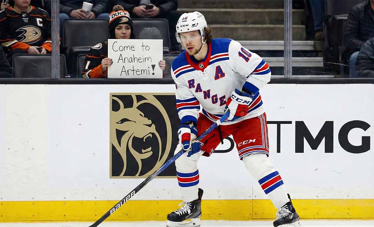 Artemi Panarin, wearing a white New York Rangers away jersey with a blue 'A', is on the ice with his stick during a game. In the background, a fan holds a sign that reads, "Come to Anaheim Artemi!".
