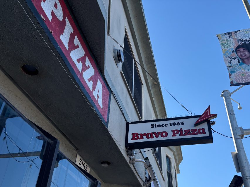 A street view of the Bravo Pizza storefront, featuring signs reading “PIZZA” and “Bravo Pizza Since 1963,” with a building and a hanging banner in the background.