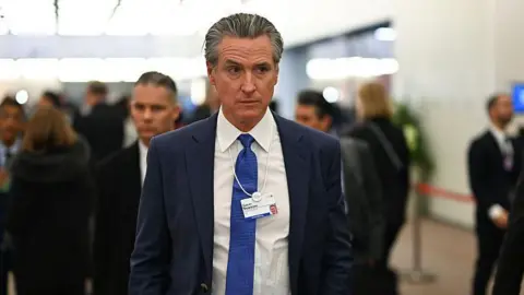 Bloomberg via Getty Images California Gov Gavin Newsom is seen walking through a hallway at the Davos World Economic Forum. He has on a suit with a blue tie and has on a lanyard identifying himself. 