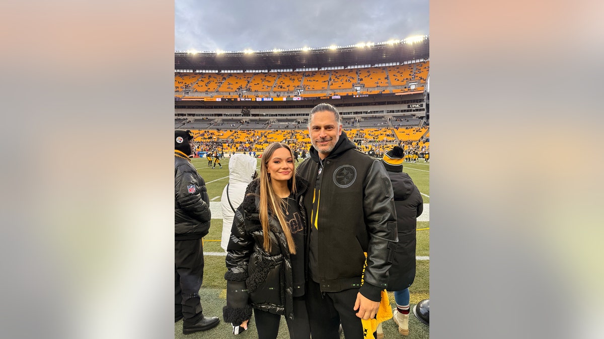 Joe Manganiello and Caitlin O'Connor smiling and wearing matching leather jackets standing on a football field.