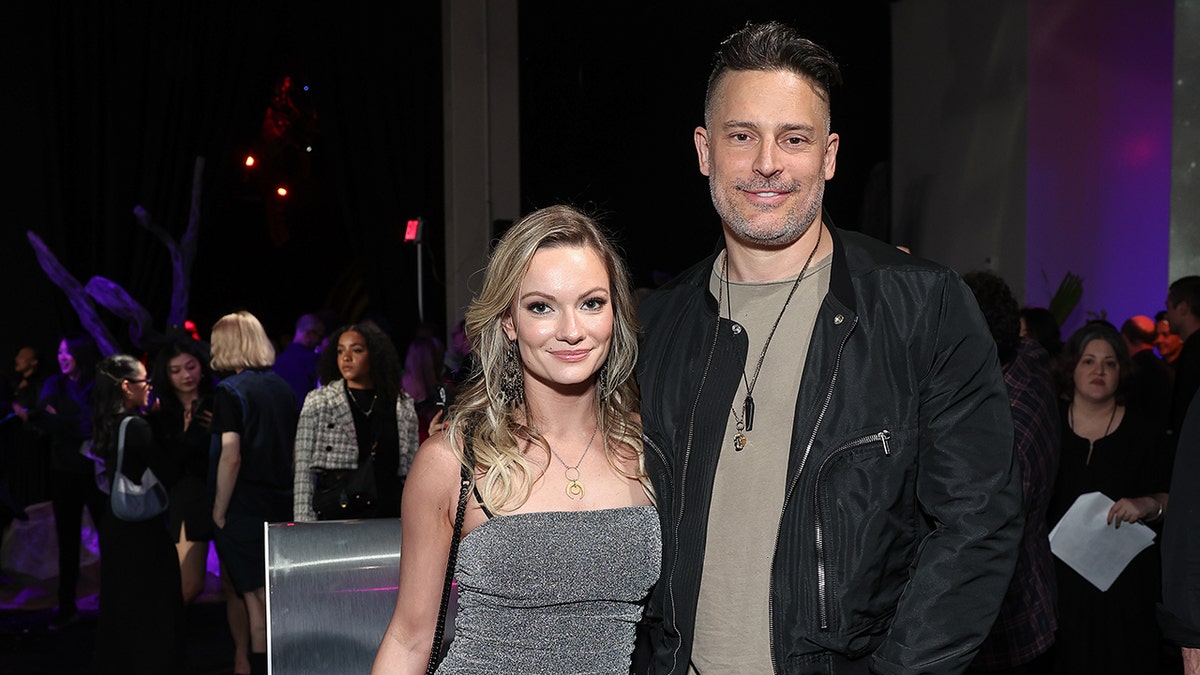 Caitlin O'Connor wearing a sleeveless grey dress standing next to Joe Manganiello wearing a black jacket and a grey shirt.