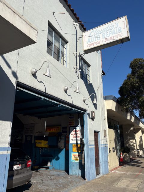 A blue and white building with an open garage labeled "Claudio's Auto Repair Foreign & Domestic." A car is parked inside and signs are posted by the entrance.