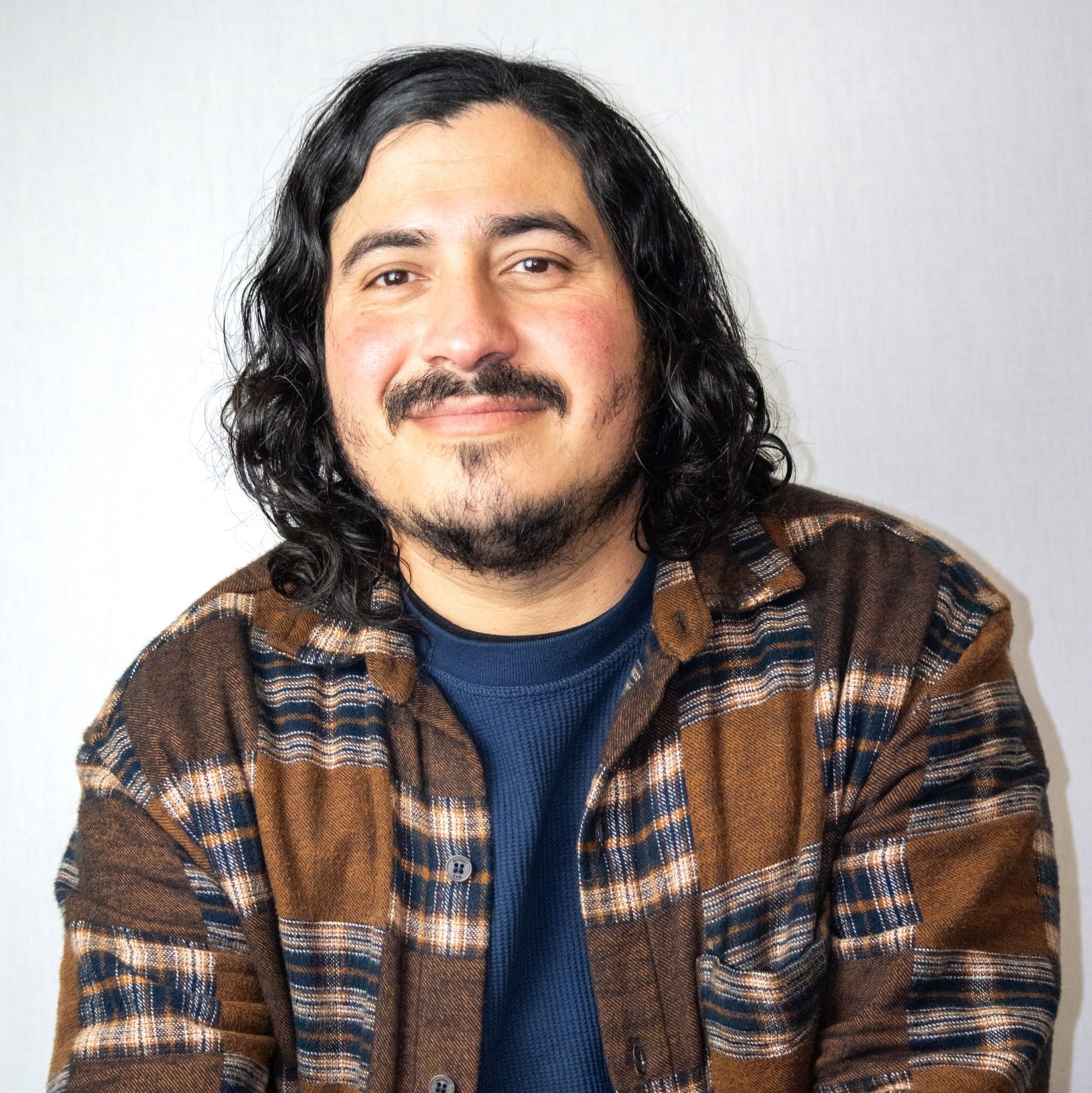 A man with medium-length curly hair and a beard smiles at the camera, wearing a brown plaid shirt over a navy shirt, in front of a plain light-colored background.