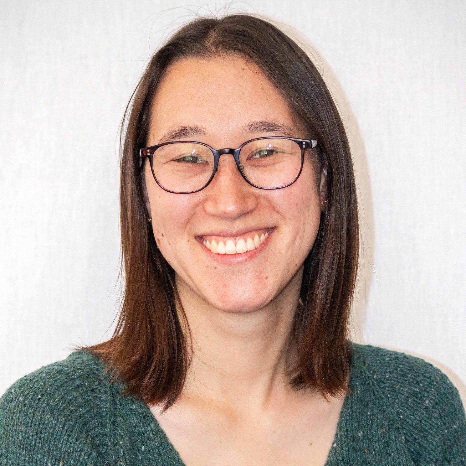 A person with straight brown hair, wearing glasses and a green sweater, smiles at the camera in front of a plain light background.