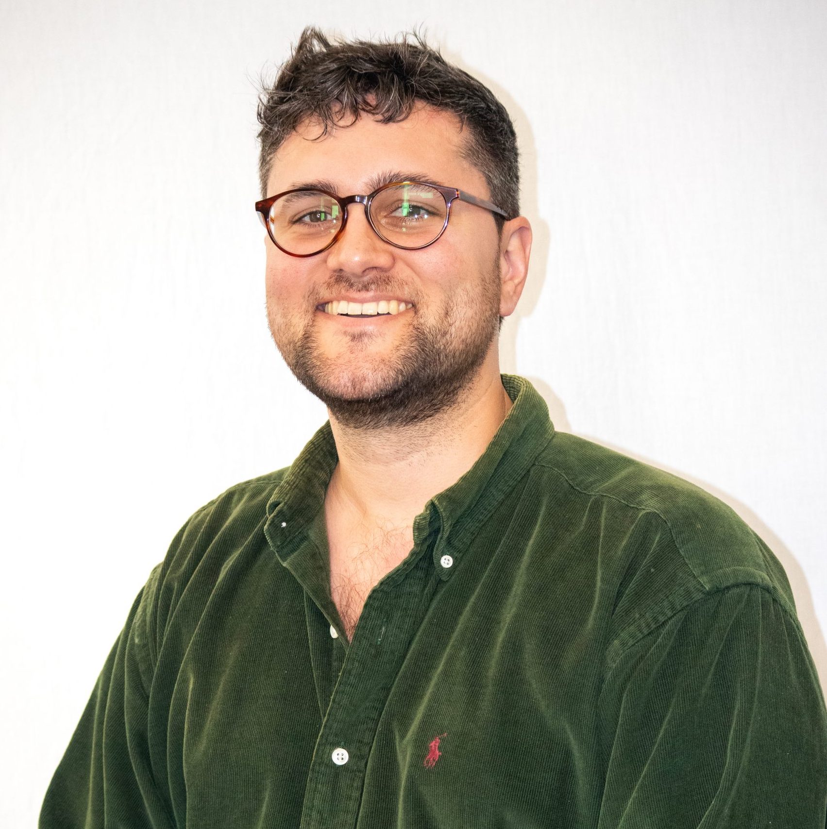 Man with short dark hair, glasses, and a beard, wearing a green button-up shirt, smiling in front of a plain white background.