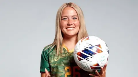 Julia Lucero A woman with blonde straight hair standing in front of a white background. She is in a green and gold football top with the number 26 on it. She is holding a football and smiling. She has dark green shorts and long socks on, with bronze and black football boots