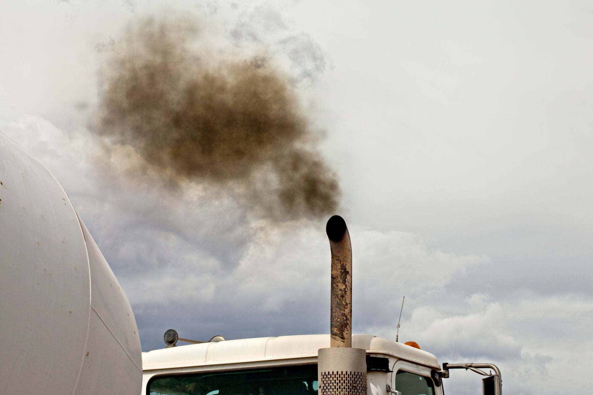 A white dump truck spews black exhaust smoke into a blue sky. Only the top of the truck is visible with the sky taking up half the image.