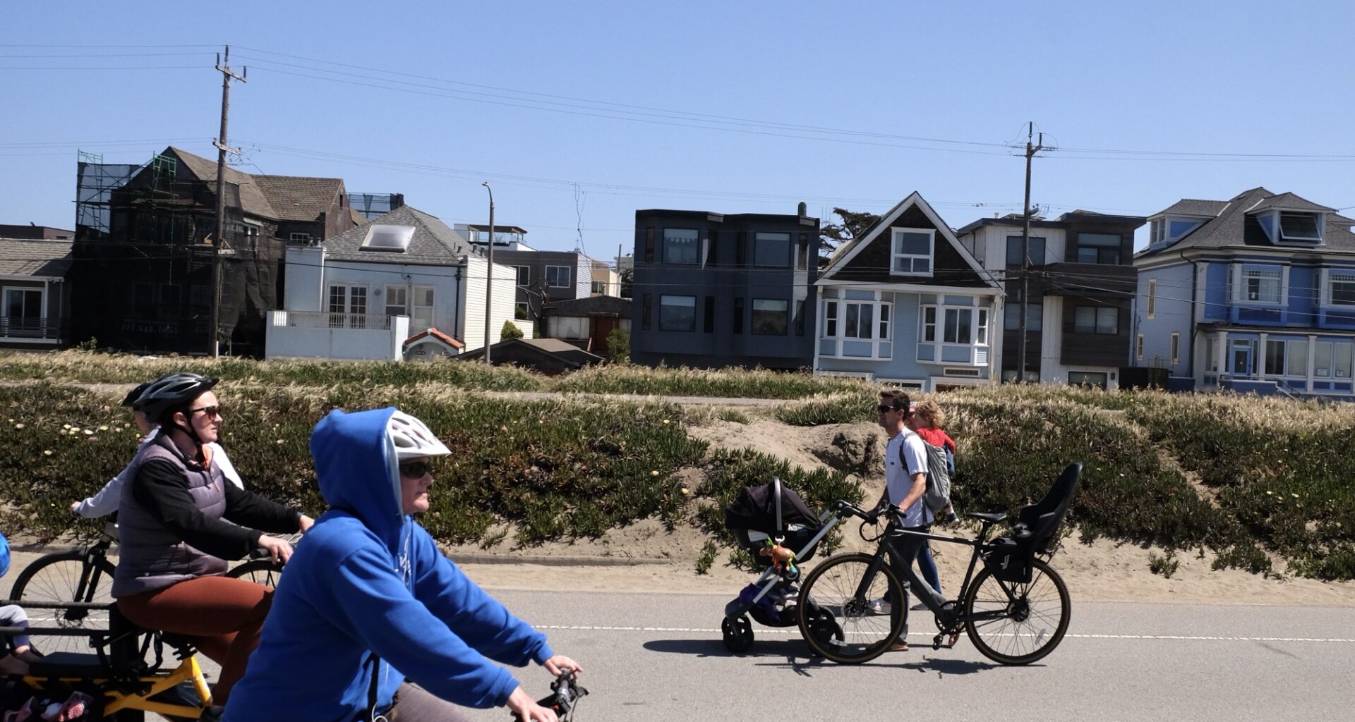 People riding bicycles along a road near residential houses on a clear day. Some cyclists wear helmets. A stroller is attached to one bicycle.