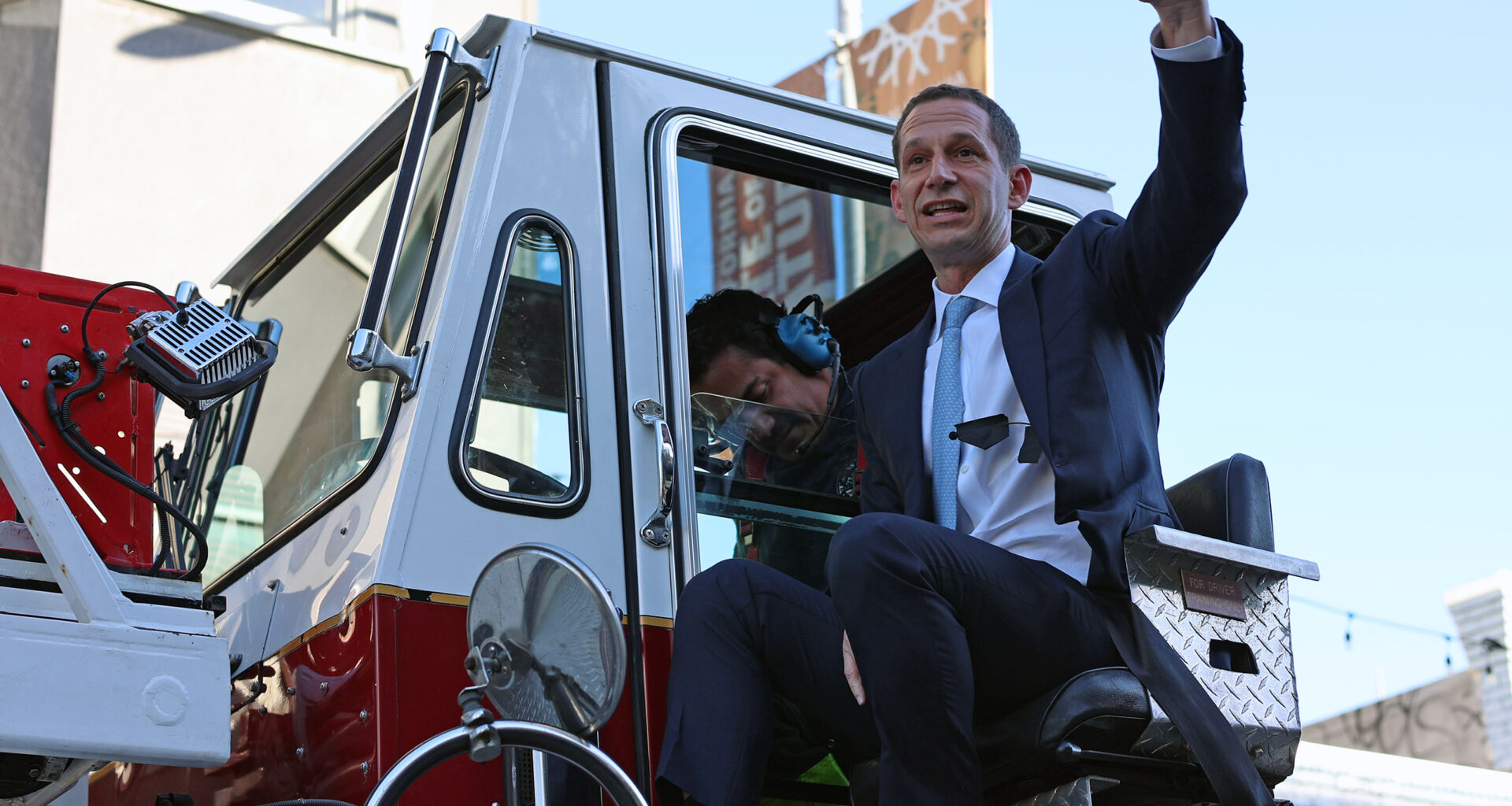 A man in a suit sits on a fire truck, gesturing enthusiastically, while another person is seated inside. The background includes buildings and a streetlight.