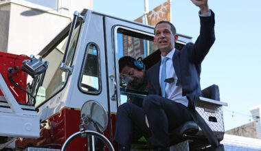 A man in a suit sits on a fire truck, gesturing enthusiastically, while another person is seated inside. The background includes buildings and a streetlight.