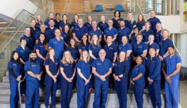 Dozens of emergency medicine team members in matching blue scrubs stand on a staircase inside a modern building.