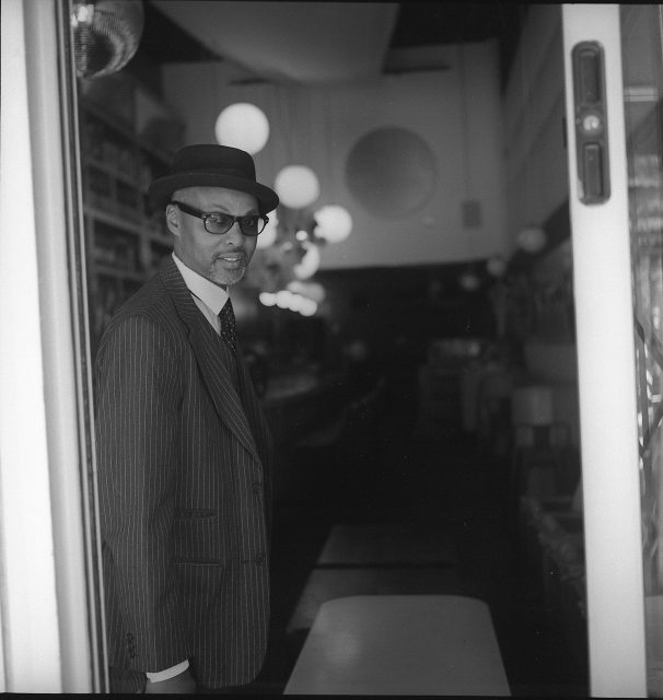 A man in a pinstripe suit, tie, and hat stands at the entrance of a dimly lit establishment with round lights visible in the background.
