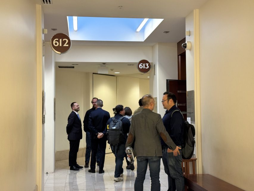A group of people stand and talk in a hallway outside rooms 612 and 613, under a skylight, with beige walls and security cameras visible.