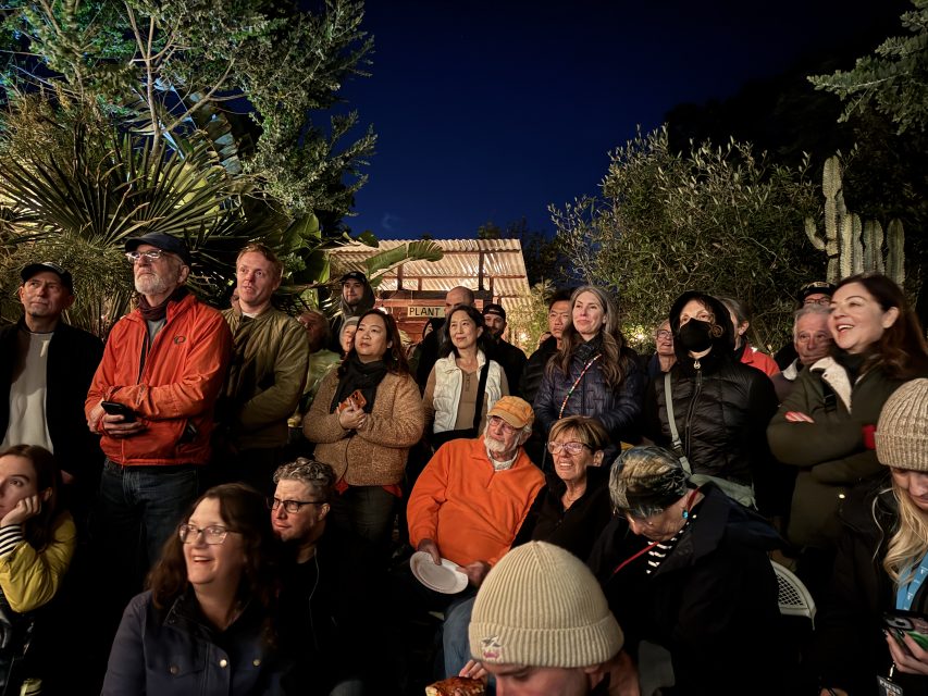 A group of people, some seated and some standing, gather outdoors at night, dressed in warm clothing, under trees and near a building with a tile roof.