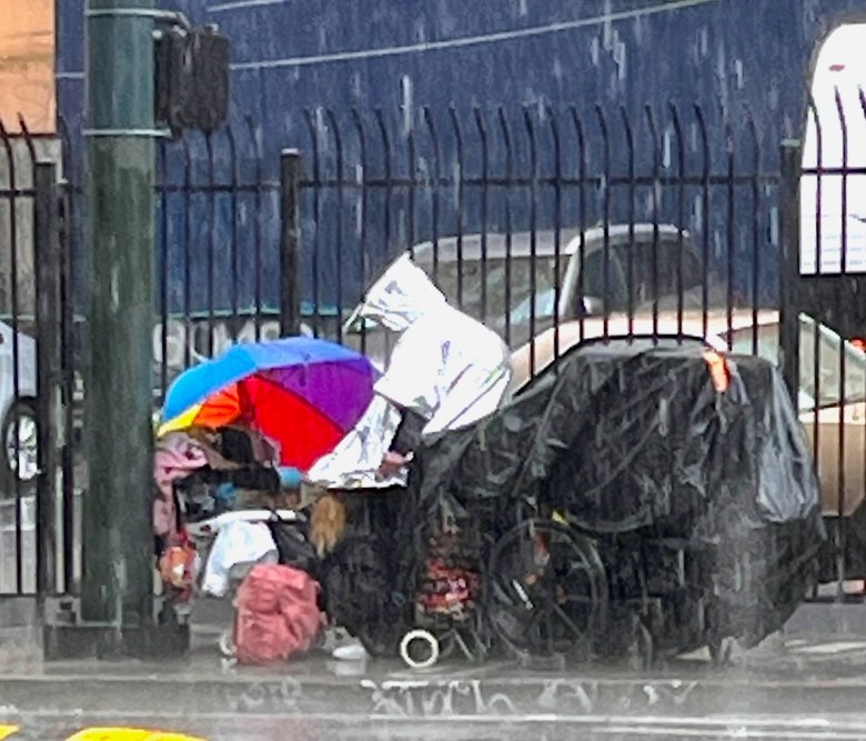 A person in a silver rain hood in the rain on a street corner amidst a pile of belongings.