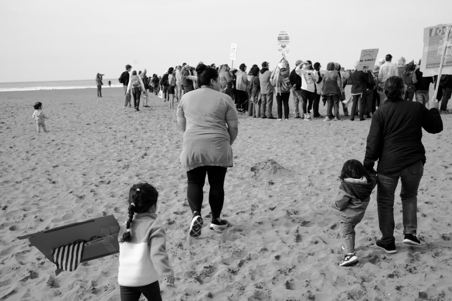 A group of people gathered on a sandy beach, some holding signs, while a few children and adults walk toward the crowd.