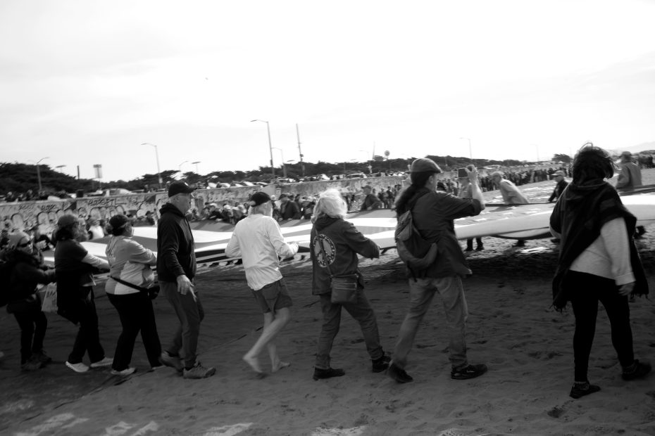A group of people walk together on sand, carrying a large object, with a crowd and graffiti-covered wall in the background.