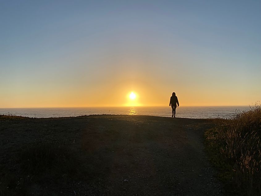 A woman stands atop a hill, shrouded in shadow as the sun sets over the ocean in the distance.