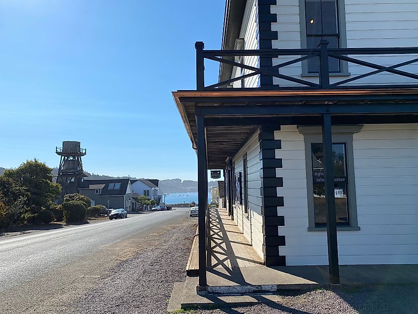 A two-story, old fashioned building stands before a wooden water tower. A bluebird sky spans the water in the distance.