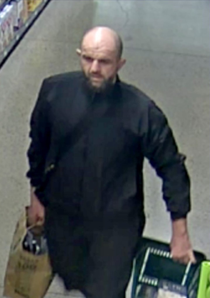 Surveillance image of a bald man with a beard, wearing a black shirt, carrying a brown paper bag and a green plastic tote.