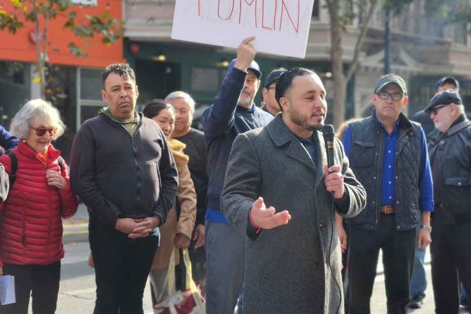A man speaks into a microphone at an outdoor gathering. People stand around him, including a person holding a sign in the background. Trees and buildings are visible.