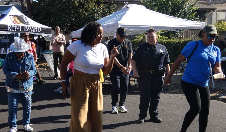 Five people, one of them in a police uniform, dance on a street in the sunshine