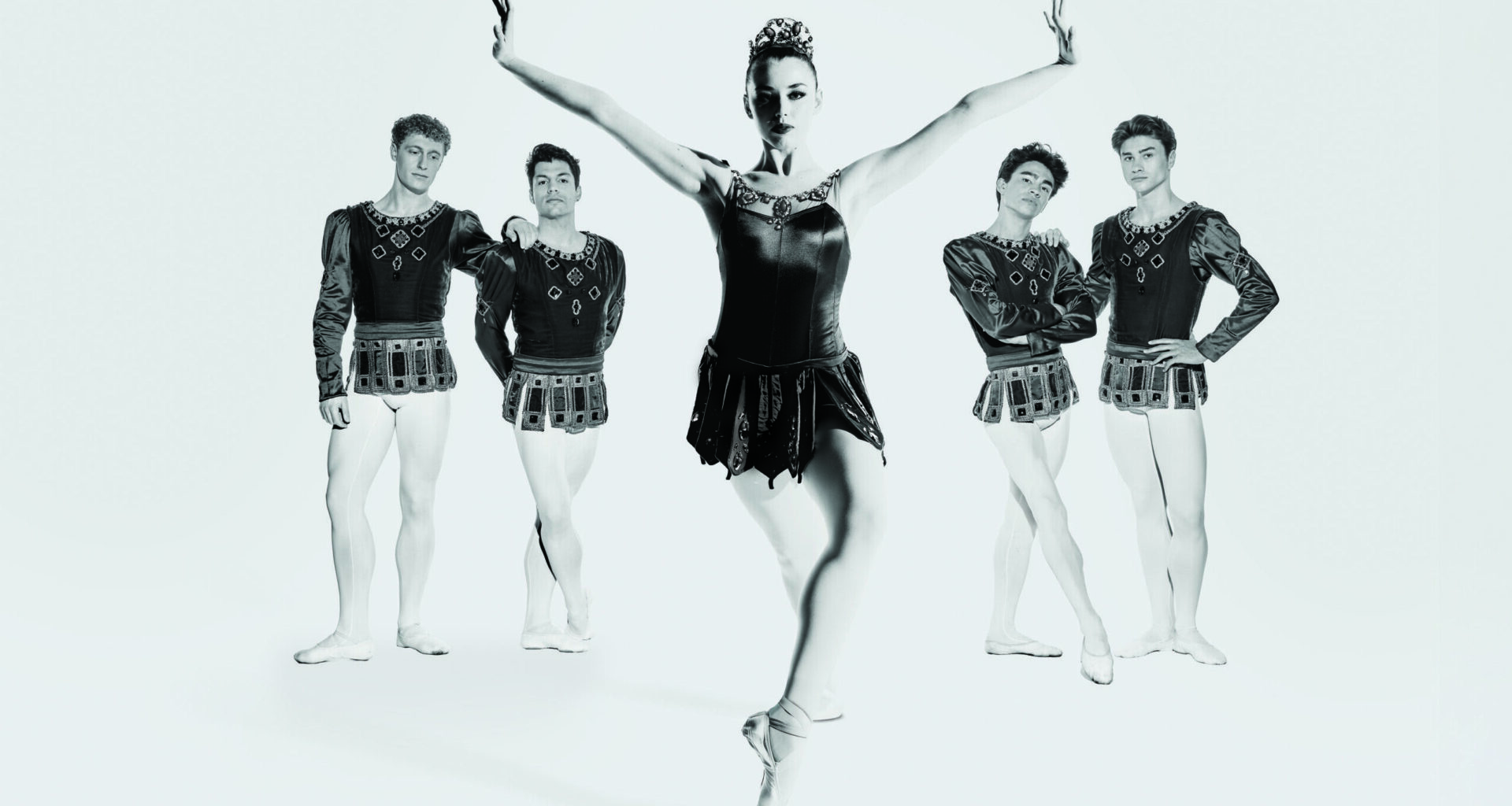A silver-tone image of five dancers in "Rubies," with four men standing casually in the back as a woman, in center, leans forward with one foot propped on pointe. She flexed her hands and reaches her arms up on a "V" shape as she looks into the camera.