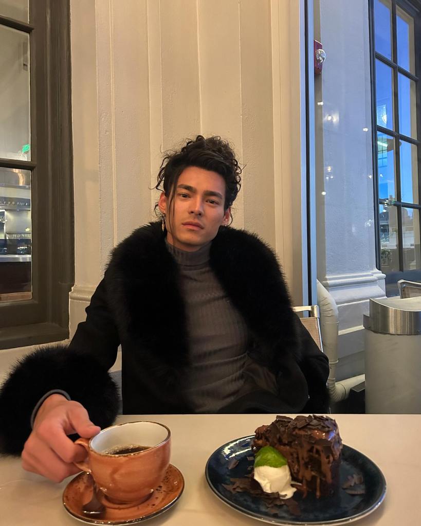 Miguel Marchese in a dark fur coat holding a cup of coffee with a chocolate cake on the table.