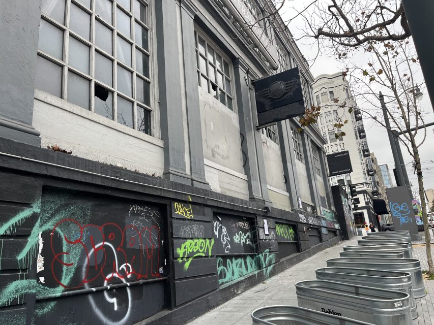 Gray building with broken windows and graffiti on lower walls, leafless tree in front, metal bins lined up along the sidewalk on a cloudy day.
