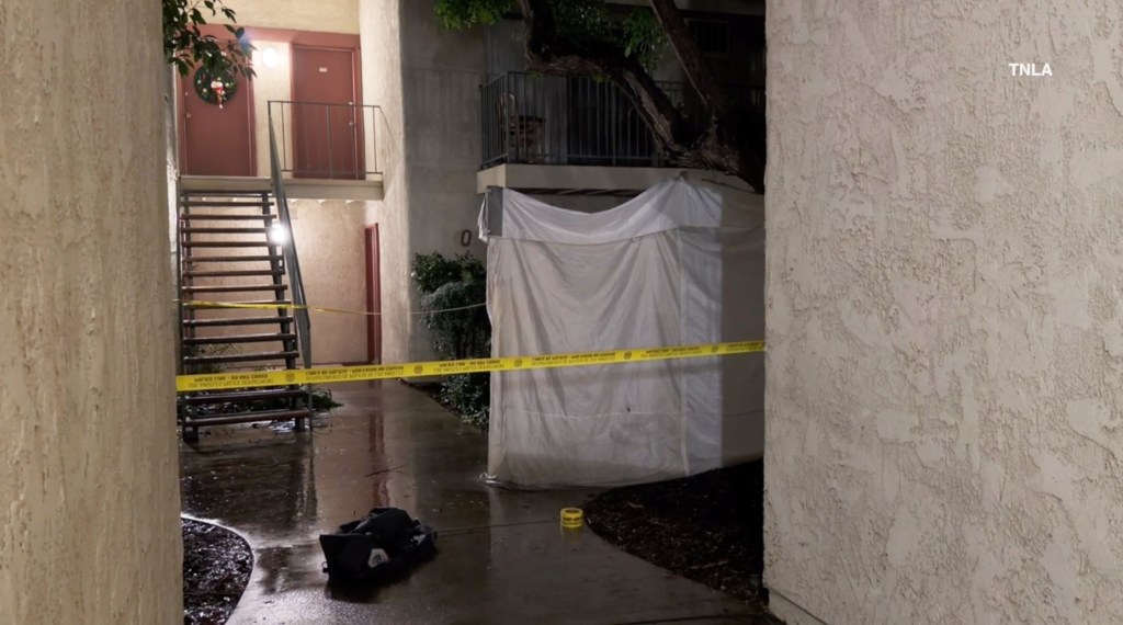 A view of an apartment complex hallway at night, with yellow crime scene tape cordoning off an area near a metal staircase and a white tarp covering a section of the building.