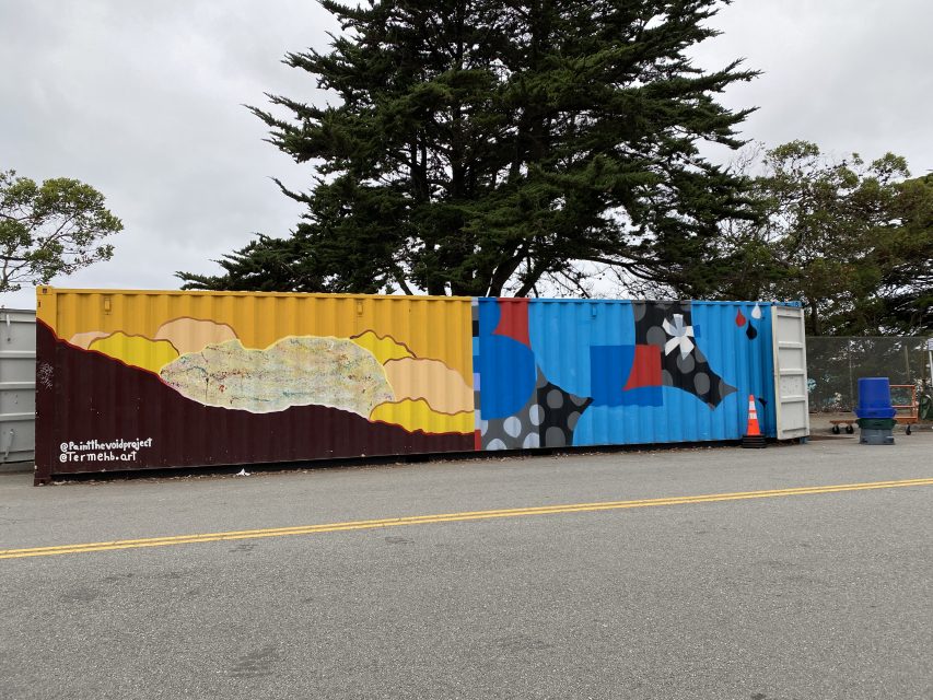 A large shipping container painted with abstract art in yellow, red, blue, and black stands on a paved area, with trees and a cloudy sky in the background.