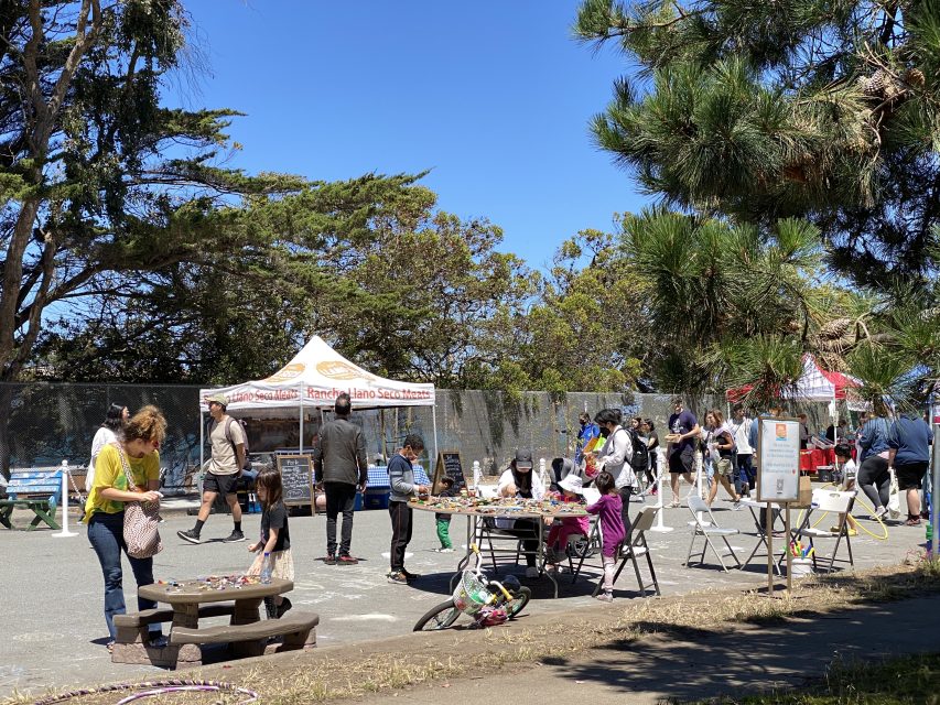 People visit outdoor craft and food stalls on a sunny day, with children and adults gathered around tables and a tree in the foreground.