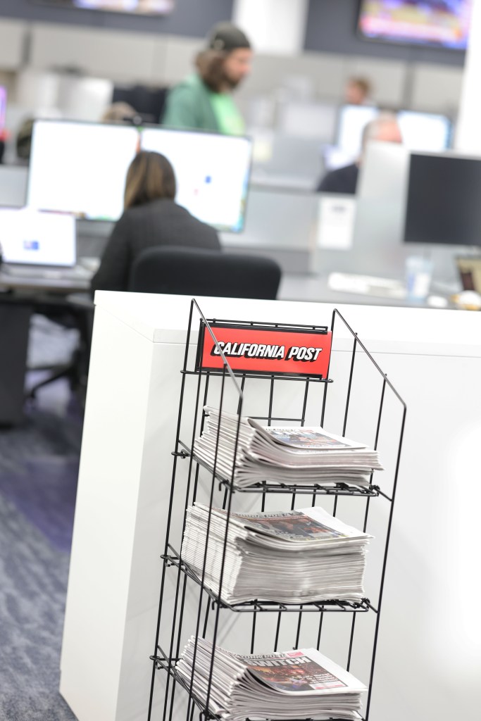 Newspapers stacked in a display stand with a "California Post" sign, in an office setting.