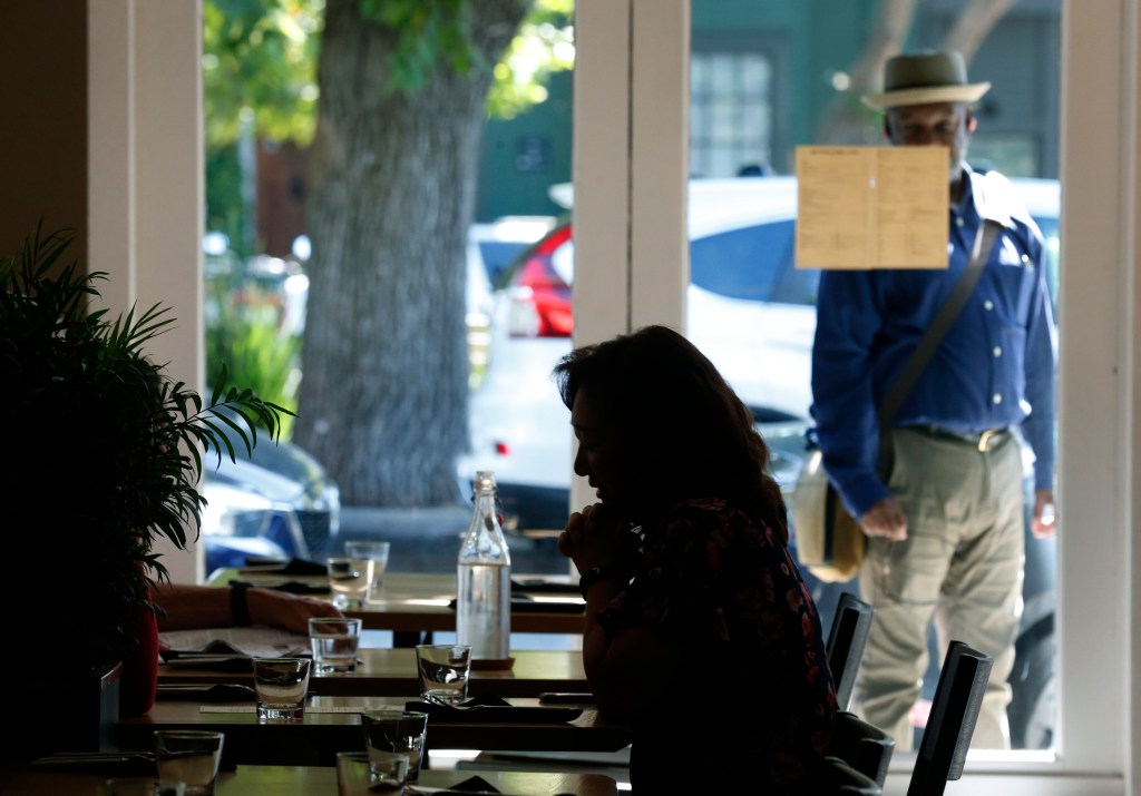 A woman at a cafe table while a man outside looks at the menu posted on the window.