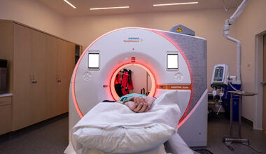 Large white cylindrical CT scanner with orange light and patient’s hands crossed in the foreground.