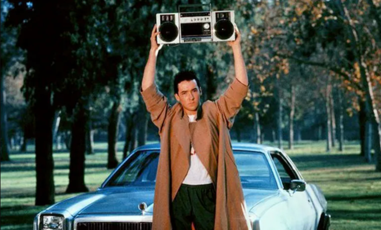 A man in a trench coat standing in front of a car and holding a boom box.