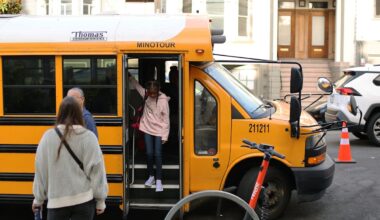 A child steps off a yellow minibus while two adults wait nearby on a city street with parked cars, an orange traffic cone, and a scooter in the foreground.