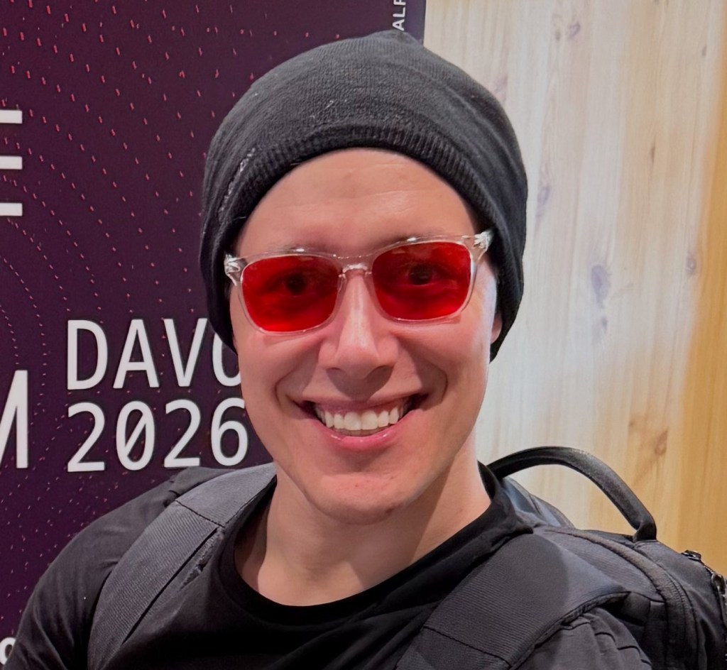 Sebastian Heyneman, smiling and wearing red sunglasses and a black beanie, against a purple backdrop with "DAVOS 2026" visible.