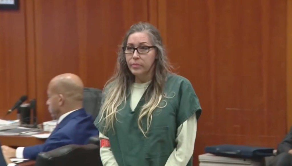 Shannon O'Connor in a courtroom, wearing a green jail uniform and glasses, with another person out of focus in the background.