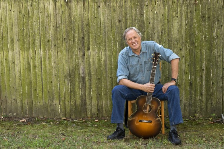 A man dressed in denim sits in front of an old fence holding a guitar by the neck.